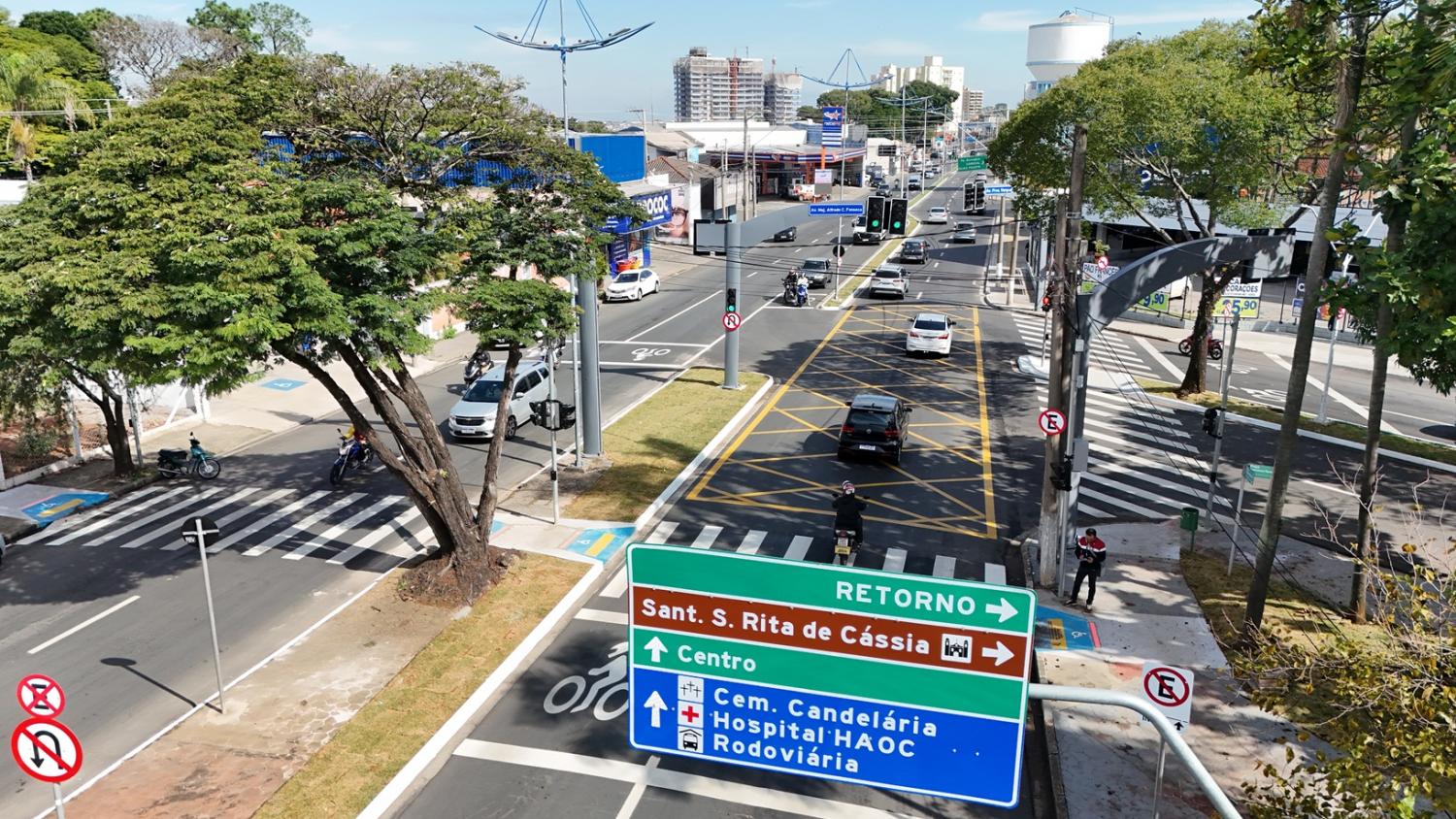 Fotografia aérea capturada por drone mostrando a vista em perspectiva do cruzamento e fluxo de veículos na Avenida Presidente Vargas sob iluminação solar diurna. Em primeiro plano, no canto inferior central e direito, destaca-se uma grande placa indicativa de trânsito em pórtico metálico com orientações nas cores verde, marrom e azul. O cruzamento possui sinalização horizontal completa, incluindo uma ampla caixa amarela de marcação de área confluente (não feche o cruzamento) no centro da pista, faixas de pedestres zebradas e linhas de retenção. Em ambas as pistas, há movimentação de carros, motos e um pedestre caminhando pela calçada à direita. Fotografia aérea capturada por drone mostrando a vista em perspectiva do cruzamento e fluxo de veículos na Avenida Presidente Vargas sob iluminação solar diurna. Em primeiro plano, no canto inferior central e direito, destaca-se uma grande placa indicativa de trânsito em pórtico metálico com orientações nas cores verde, marrom e azul. O cruzamento possui sinalização horizontal completa, incluindo uma ampla caixa amarela de marcação de área confluente (não feche o cruzamento) no centro da pista, faixas de pedestres zebradas e linhas de retenção. Em ambas as pistas, há movimentação de carros, motos e um pedestre caminhando pela calçada à direita.
